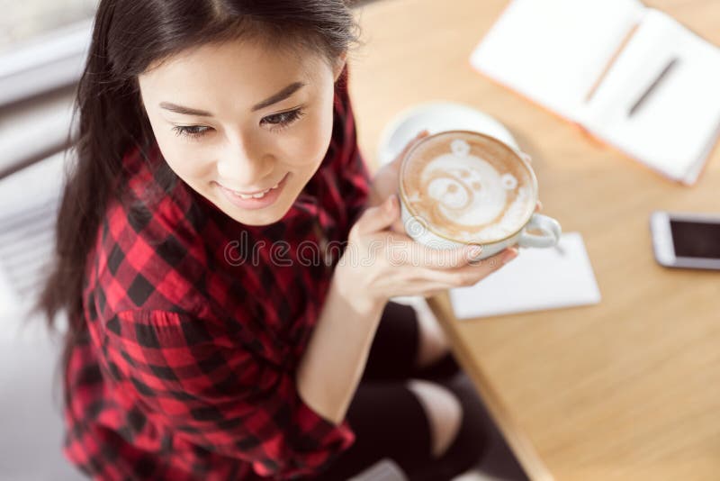 Mulher moreno nova na camisa quadriculado que guarda o copo branco e que bebe o café do cappuccino com urso decorativo foto de stock Mulher moreno nova na camisa quadriculado que guarda o copo branco e que bebe o café do cappuccino com urso decorativo foto de stock