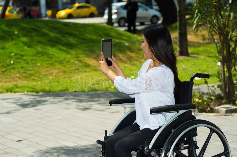 Mulher moreno nova que senta-se na cadeira de rodas que sorri com atitude positiva, usando o telefone celular, fora ambiente imagem de stock royalty free Mulher moreno nova que senta-se na cadeira de rodas que sorri com atitude positiva, usando o telefone celular, fora ambiente imagem de stock royalty free