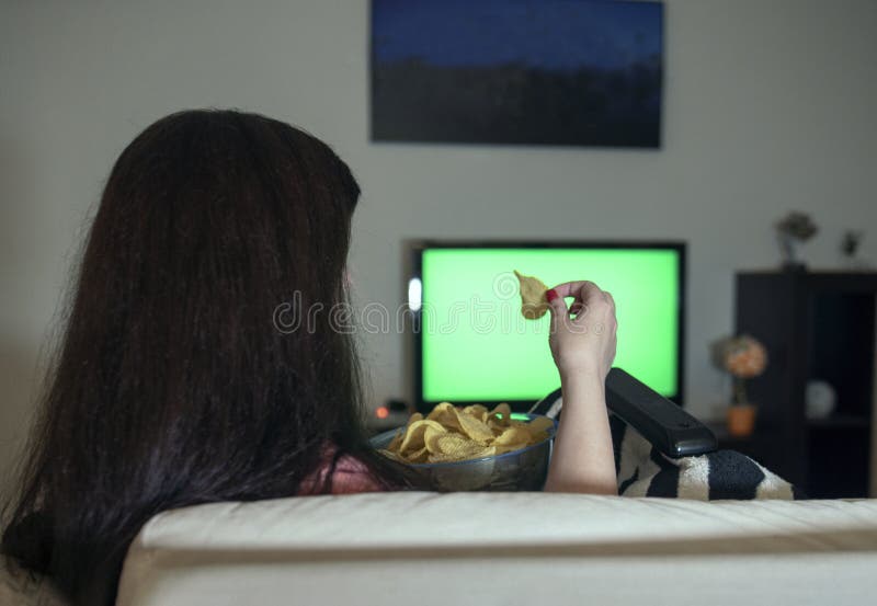 Mulher moreno que senta em casa a noite de relaxamento que come microplaquetas de batata e que olha a televisão, uma tela verde fotografia de stock royalty free Mulher moreno que senta em casa a noite de relaxamento que come microplaquetas de batata e que olha a televisão, uma tela verde fotografia de stock royalty free