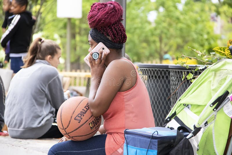 A mulher negra com tranças vermelhas senta-se no local de reunião que guarda um basquetebol e que fala em um telefone cor-de-rosa foto de stock A mulher negra com tranças vermelhas senta-se no local de reunião que guarda um basquetebol e que fala em um telefone cor-de-rosa foto de stock