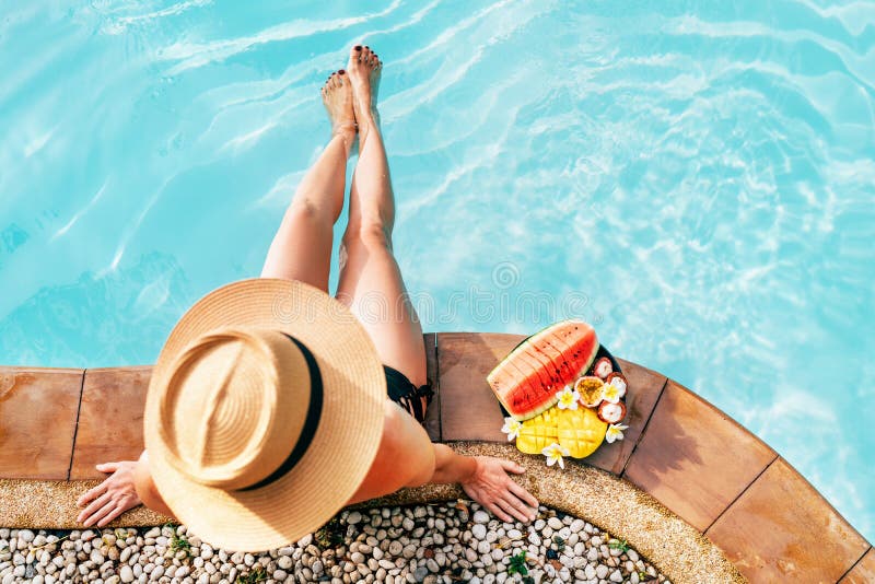 Mulher no chapéu de palha que senta-se no lado da piscina com a placa da opinião superior da câmera dos frutos tropicais