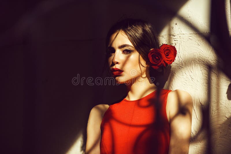 Woman in red dress and roses in hair standing at wall. Young fashionable model in shadow on sunny day. Fashion, beauty salon. Woman in red dress and roses in hair standing at wall. Young fashionable model in shadow on sunny day. Fashion, beauty salon