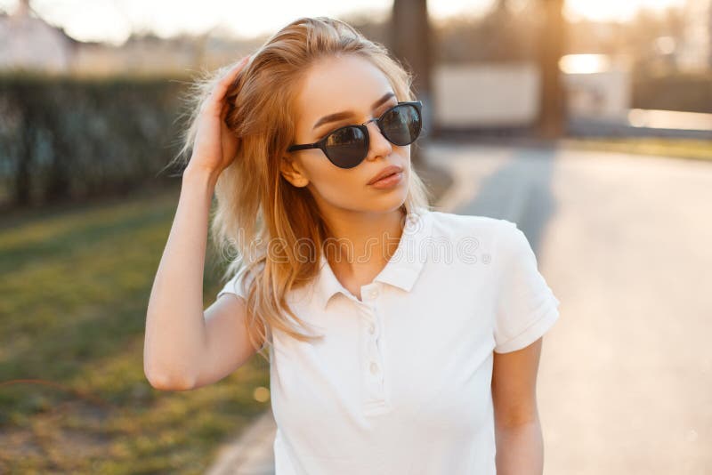A mulher nova à moda moderna do moderno no t-shirt branco na moda do polo em óculos de sol pretos está e levantar fotografia de stock A mulher nova à moda moderna do moderno no t-shirt branco na moda do polo em óculos de sol pretos está e levantar fotografia de stock
