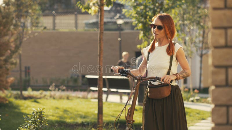 Mulher que anda ao lado da bicicleta da equitação no parque da cidade Parque da bicicleta da mulher imagem de stock royalty free Mulher que anda ao lado da bicicleta da equitação no parque da cidade Parque da bicicleta da mulher imagem de stock royalty free