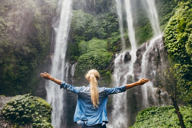 Rear view of young woman standing in front of waterfall with her hands raised. Female tourist with her arms outstretched looking at waterfall. Rear view of young woman standing in front of waterfall with her hands raised. Female tourist with her arms outstretched looking at waterfall.