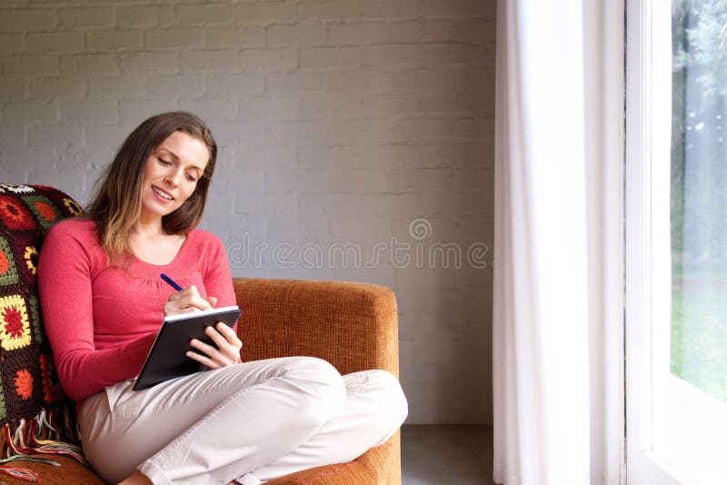 Mulher que senta-se no sofá em casa e que escreve no livro imagem de stock royalty free Mulher que senta-se no sofá em casa e que escreve no livro imagem de stock royalty free