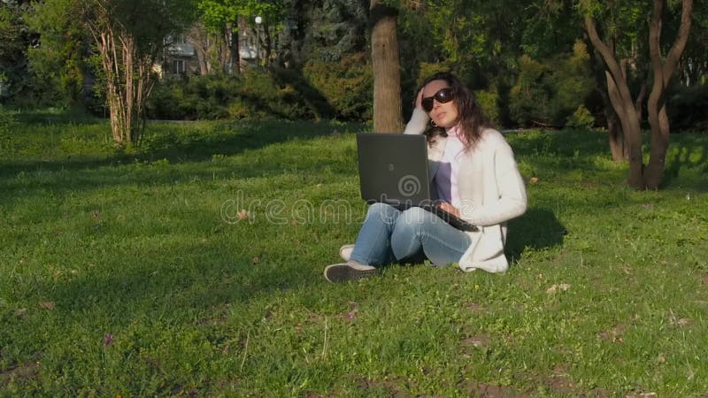 Mulher que trabalha no portátil na natureza A menina no parque está sentando-se na grama com o computador Jovem mulher no worki d