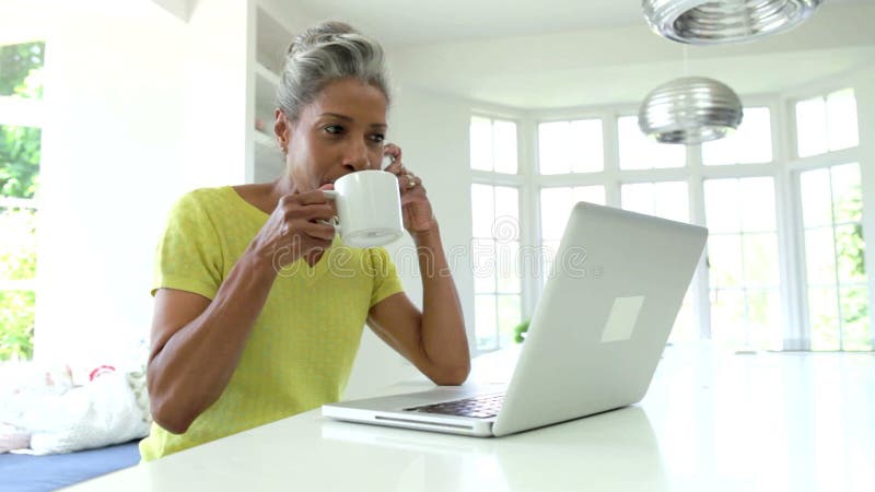 Mulher que usa o portátil e falando no telefone na cozinha em casa