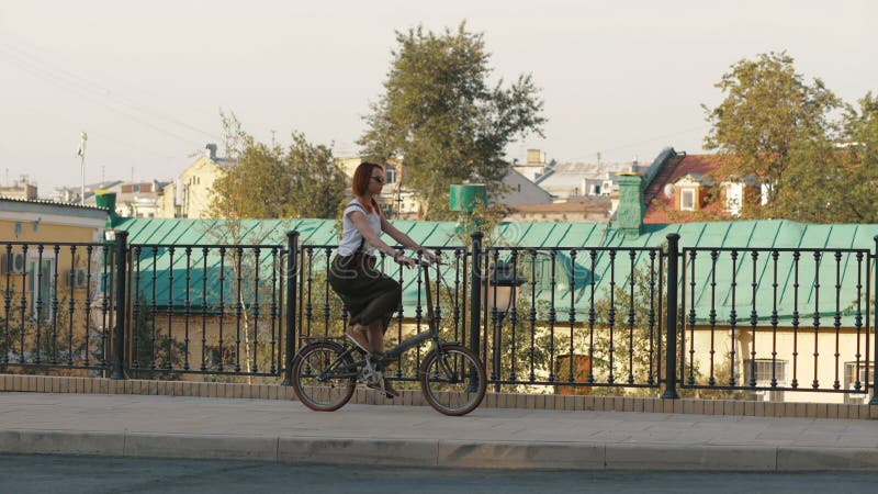 Mulher Redhaired que monta uma bicicleta na cidade em construções do fundo e em casa do telhado fotografia de stock Mulher Redhaired que monta uma bicicleta na cidade em construções do fundo e em casa do telhado fotografia de stock
