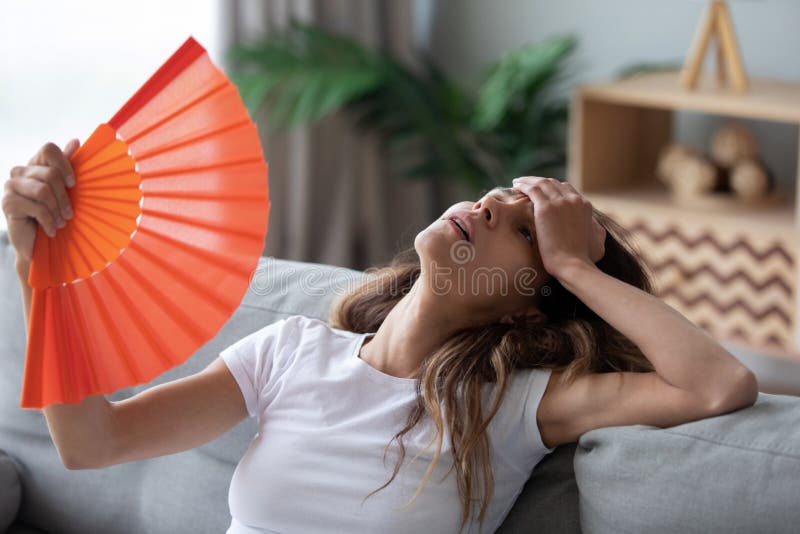 Overheated woman sitting on couch, waving orange paper fan close up, girl feeling unwell, suffering from heating at home, feeling discomfort, hot summer weather or fever, sitting on couch alone. Overheated woman sitting on couch, waving orange paper fan close up, girl feeling unwell, suffering from heating at home, feeling discomfort, hot summer weather or fever, sitting on couch alone