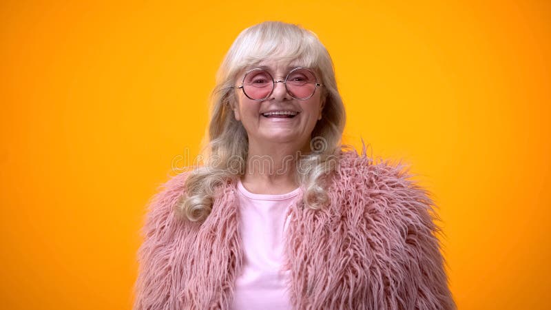 Mulher superior alegre no revestimento cor-de-rosa e em óculos de sol redondos que sorri na câmera imagem de stock Mulher superior alegre no revestimento cor-de-rosa e em óculos de sol redondos que sorri na câmera imagem de stock