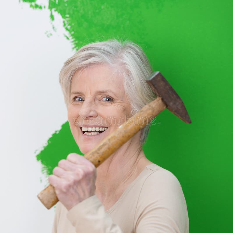 Laughing senior woman with a vivacious smile doing interior decorating at home standing with a hammer in front of a half painted green wall. Laughing senior woman with a vivacious smile doing interior decorating at home standing with a hammer in front of a half painted green wall
