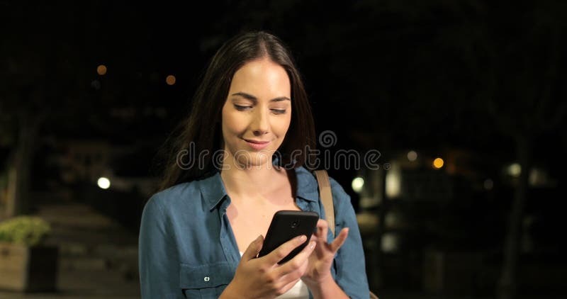 Mulher séria que anda usando o telefone esperto na noite escura