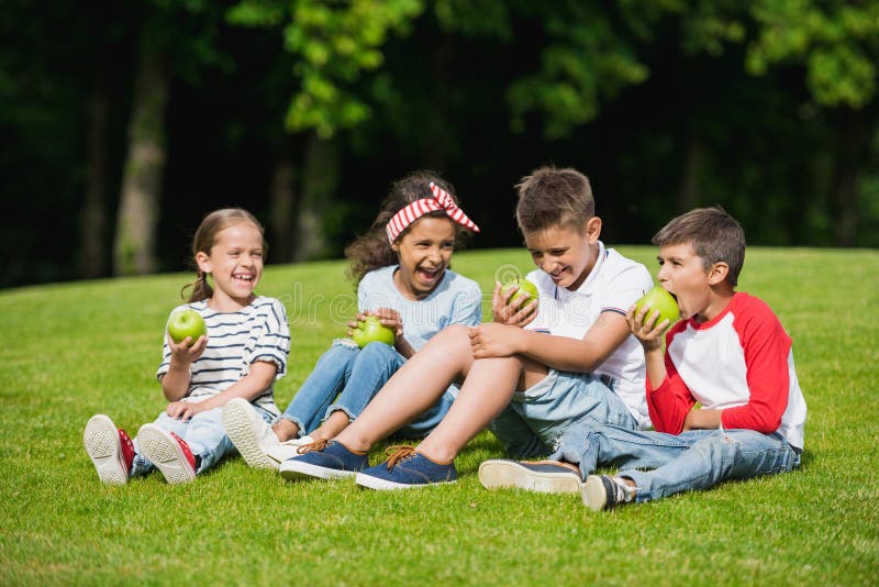 Multi-etnische kinderen die groene appelen eten terwijl het zitten samen op groen gras royalty-vrije stock afbeeldingen Multi-etnische kinderen die groene appelen eten terwijl het zitten samen op groen gras royalty-vrije stock afbeeldingen