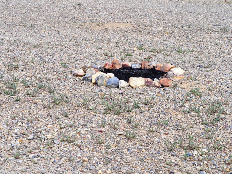 A circular arrangement of multicolored stones forms a fire ring on a gravelly terrain, surrounded by sparse vegetation. The stones vary in color, including hues of red, gray, and tan, and enclose a patch of blackened earth, indicating previous fire use. The ground is covered in small pebbles and patches of green plants, suggesting a semi-arid environment. The scene implies an outdoor camping or recreational area where fires are made safely within the boundary of the stone circle. Sparse use stock images, royalty-free photos and pictures
