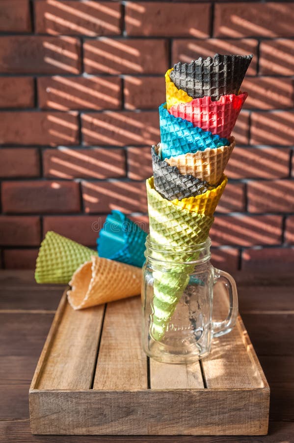 Multicolored horns for ice cream in a jar stand on a wooden box in a contrasting shadow pattern from the blinds. Red brick wall in stock photography Multicolored horns for ice cream in a jar stand on a wooden box in a contrasting shadow pattern from the blinds. Red brick wall in stock photography