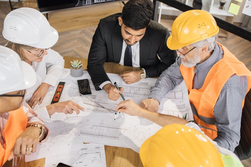 Multicultural colleagues in suits and helmets sitting at table and looking at blueprints. royalty free stock photos