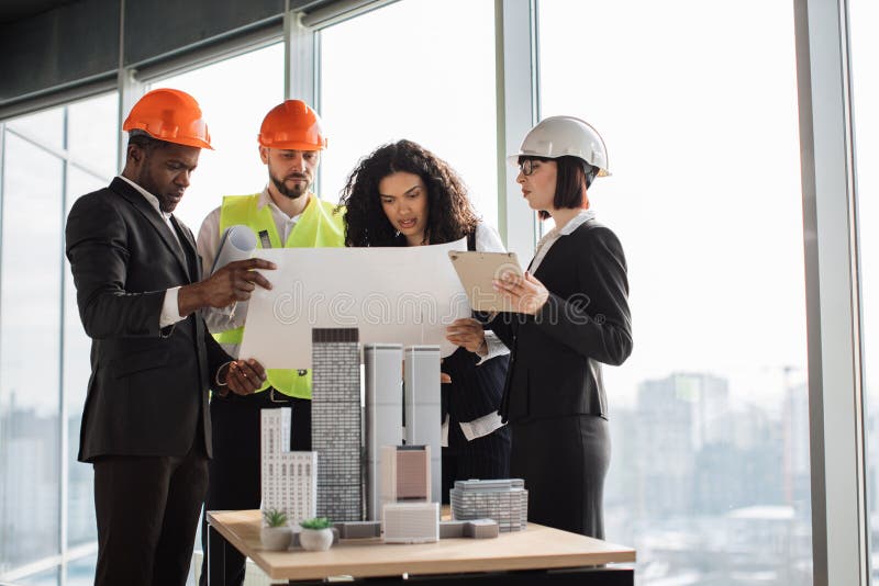 Multicultural colleagues in suits and helmets standing near table and looking at blueprint royalty free stock photography