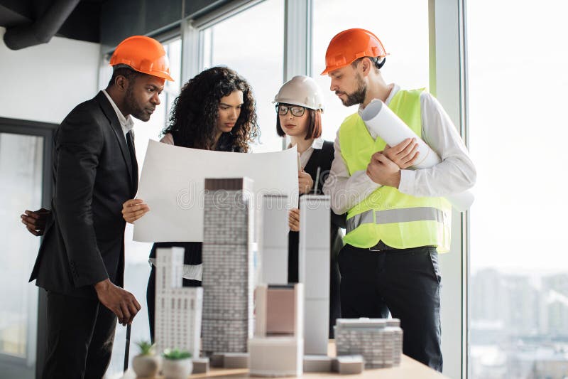 Multicultural workers in reflective vest and helmets with blueprint using city model. stock photography