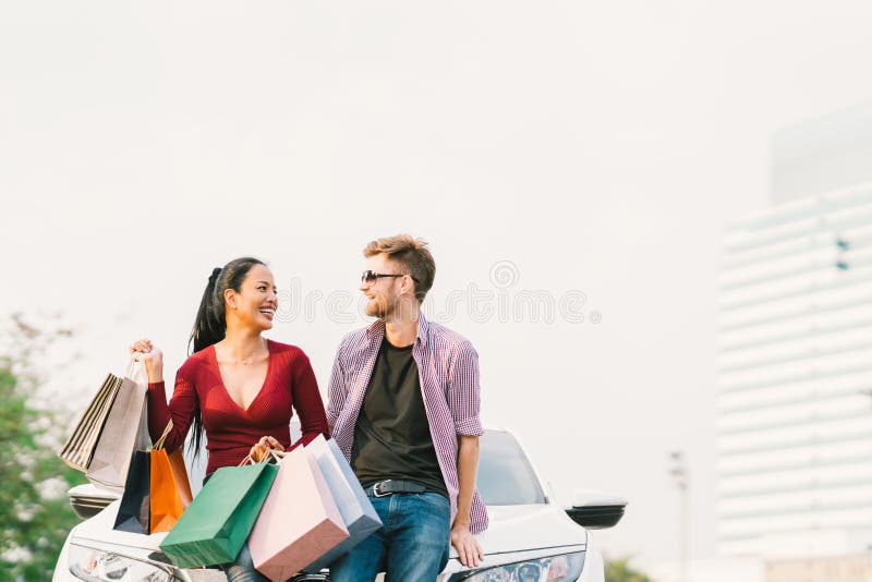 Multiethnic couple with shopping bags, smiling and sitting on white car. Love, casual lifestyle, or shopaholic concept. With copy space