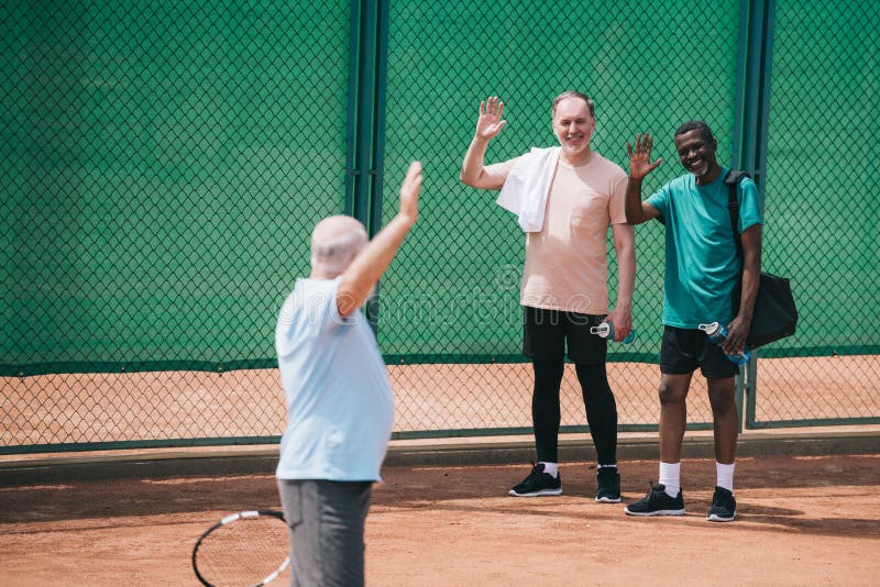 multiethnic old men greeting friend with tennis racquet