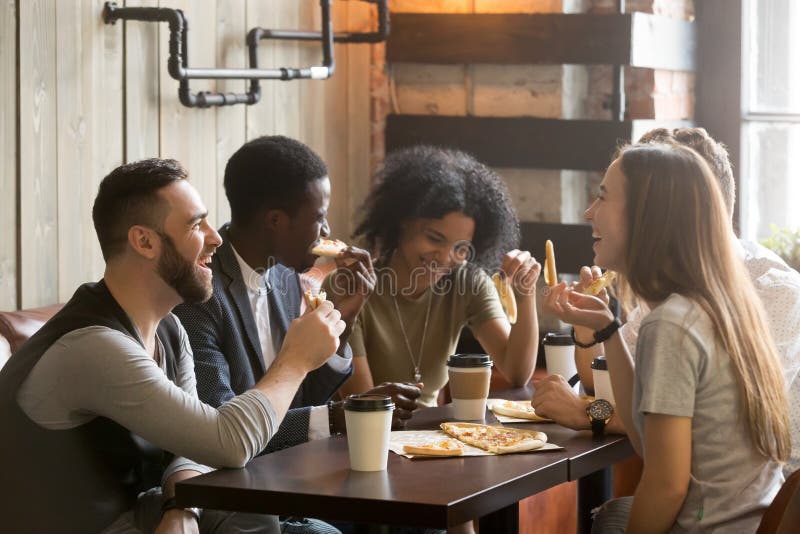 Multirazziale felici i giovani di mangiare la pizza in pizzeria, in bianco e nero allegri compagni ridendo godendo di un pasto divertirsi seduti insieme al tavolo del ristorante, diversi amici, condividere il pranzo al meeting.