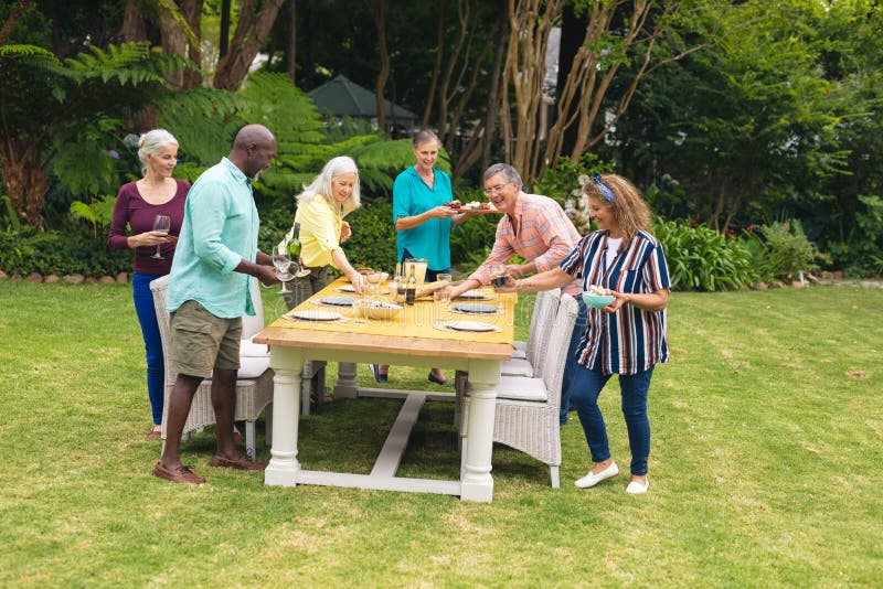Multiracial senior male and female friends arranging food on table in backyard