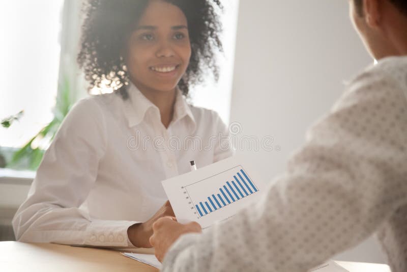 Happy multiracial millennial colleagues discussing company financial statistics at briefing in office, employees brainstorming analyzing growing sales, developing success strategies, reading reports. Happy multiracial millennial colleagues discussing company financial statistics at briefing in office, employees brainstorming analyzing growing sales, developing success strategies, reading reports