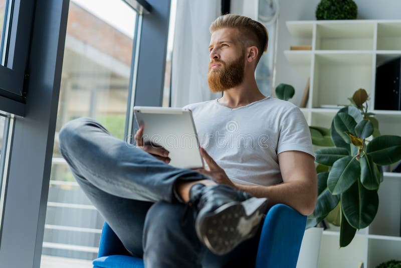 Multitasking. Handsome young man wearing glasses and working with touchpad while sitting on the couch in office royalty free stock photo Multitasking. Handsome young man wearing glasses and working with touchpad while sitting on the couch in office royalty free stock photo