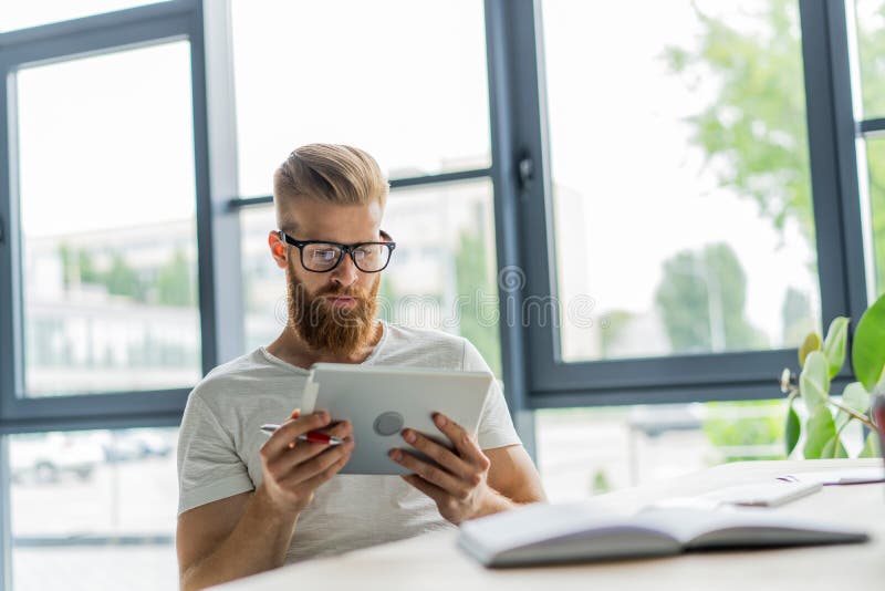 Multitasking. Handsome young man wearing glasses and working with touchpad while sitting on the couch in office royalty free stock image Multitasking. Handsome young man wearing glasses and working with touchpad while sitting on the couch in office royalty free stock image
