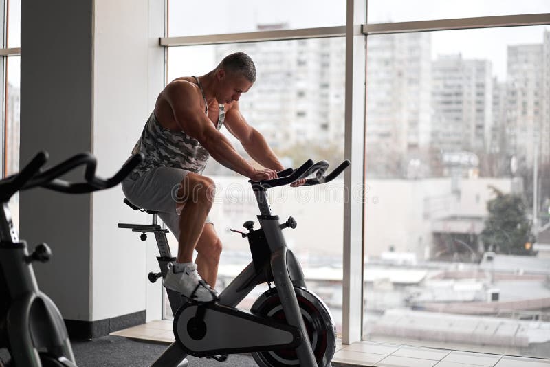 Muscular athletic bodybuilder fitness model sitting stationary bicycle in gym near big window
