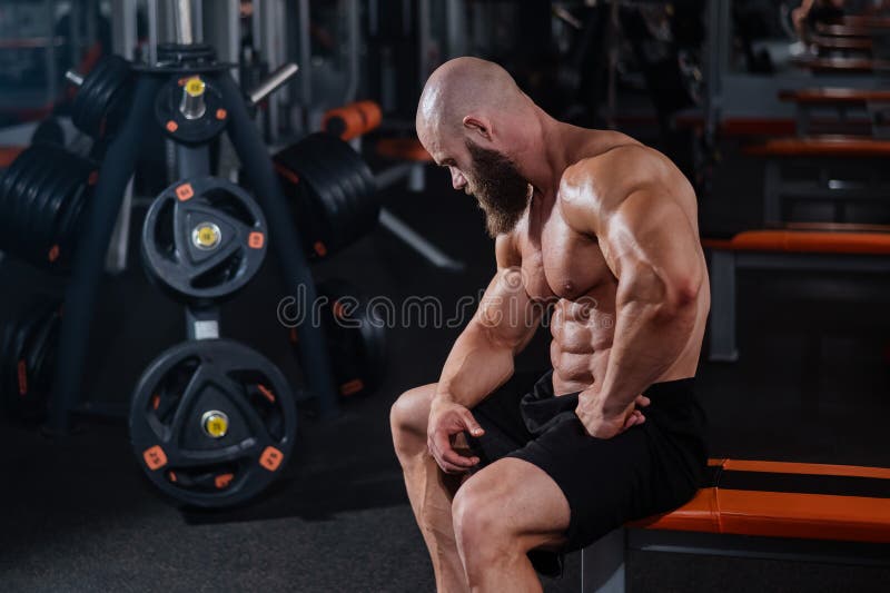 A muscular bald man in shorts is resting on a bench after a workout. Bodybuilder showing off his shape in the gym.