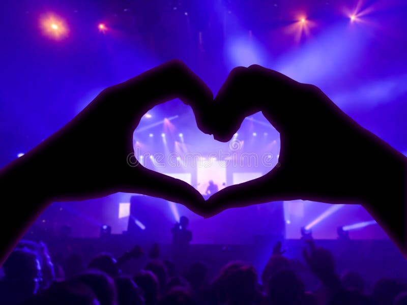 Music concert, hands raised in the shape of the heart for the music, blurred crowd and artists on stage in the background with blue spotlights