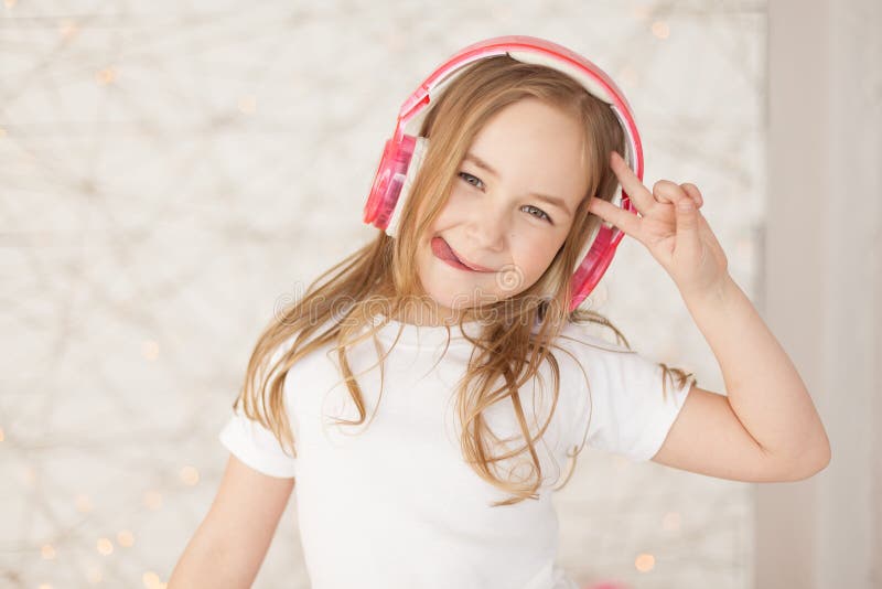 Music and technology. Young girl with pink wireless headphones make peace by hand and show tongue on background with lights. Pastel colors. indoor. Music and technology. Young girl with pink wireless headphones make peace by hand and show tongue on background with lights. Pastel colors. indoor