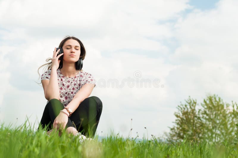 Enjoying music concept with young female sitting outside with earphones and relaxing on meadow with copyspace. Enjoying music concept with young female sitting outside with earphones and relaxing on meadow with copyspace