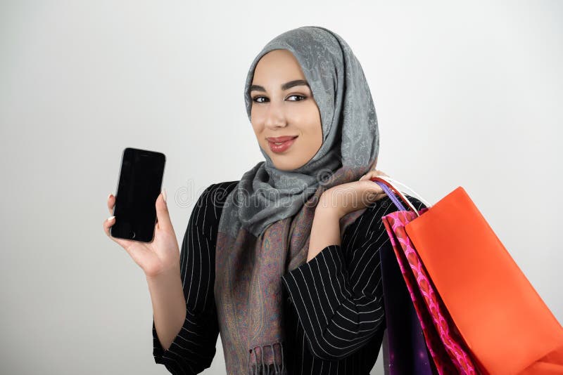 Muslim business woman wearing turban hijab headscarf showing smartphone with one hand and carrying shopping bags in stock photo Muslim business woman wearing turban hijab headscarf showing smartphone with one hand and carrying shopping bags in stock photo