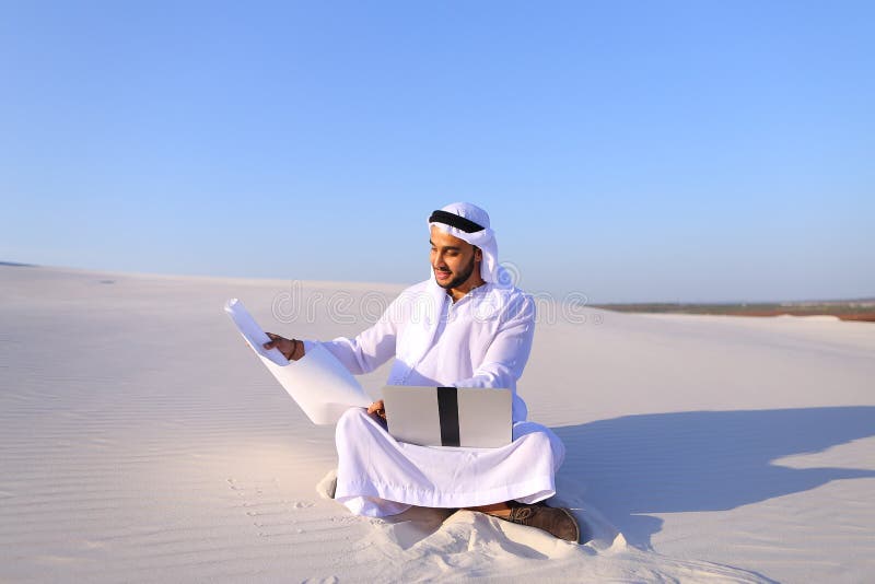 Muslim male architect sitting with laptop on sand in desert on h