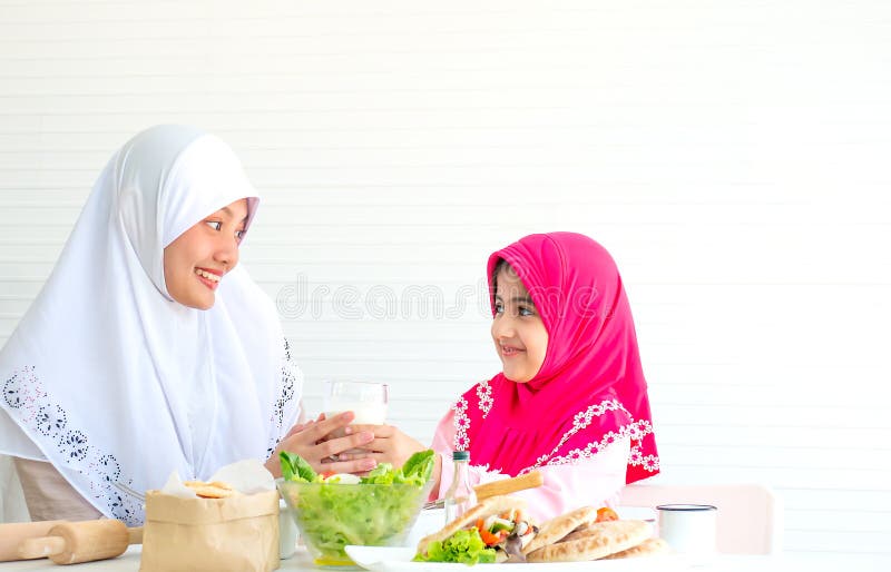 Muslim mother and little girl look to each other and smile also hold milk with bowl of vegetable salad on the table in front of stock photo Muslim mother and little girl look to each other and smile also hold milk with bowl of vegetable salad on the table in front of stock photo