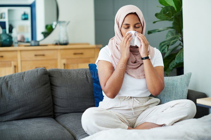 Muslim, woman and sick or sneezing on sofa in living room with illness, flu and tired at home. Female person, lounge and couch for cold, fever and virus with blowing nose on recovery or rest. Muslim, woman and sick or sneezing on sofa in living room with illness, flu and tired at home. Female person, lounge and couch for cold, fever and virus with blowing nose on recovery or rest.