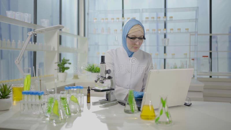 Musulmán científico del pañuelo en la cabeza en un laboratorio moderno trabaja en una laptop y un microscopio