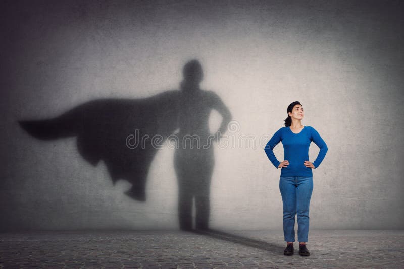 Mutige Frau hält Arme auf Hüften, lächelt zuversichtlich und wirft einen Superhelden mit Kap-Schatten an der Wand Ehrgeiz und Ges