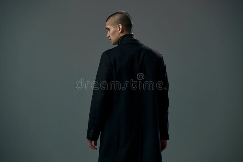 Mutiger junger Mann gekleidet im schwarzen Manteldetektiv oder -verbrecher auf grauer Studiohintergrund-Rückseitenansicht