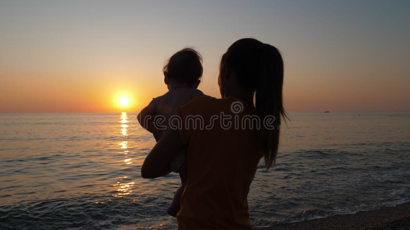 Mutter hält Kind in ihren Armen während der Betrachtung des Sonnenuntergangs durch das See- oder Ozeanufer