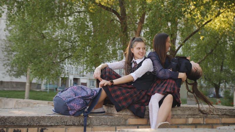 Mutter mit zwei Töchtern in der Schuluniform mit den Rucksäcken, die auf der Straße nahe der küssenden und sprechenden Schule sit