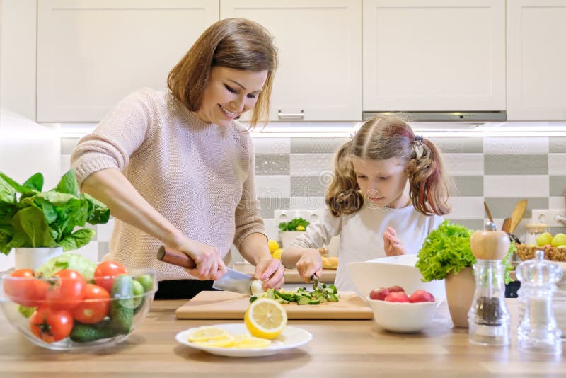 Mother and child cooking together at home in kitchen. Healthy eating, mother teaches daughter to cook, parent child communication. Woman cutting eggs, girl cutting cucumbers for salad. Mother and child cooking together at home in kitchen. Healthy eating, mother teaches daughter to cook, parent child communication. Woman cutting eggs, girl cutting cucumbers for salad