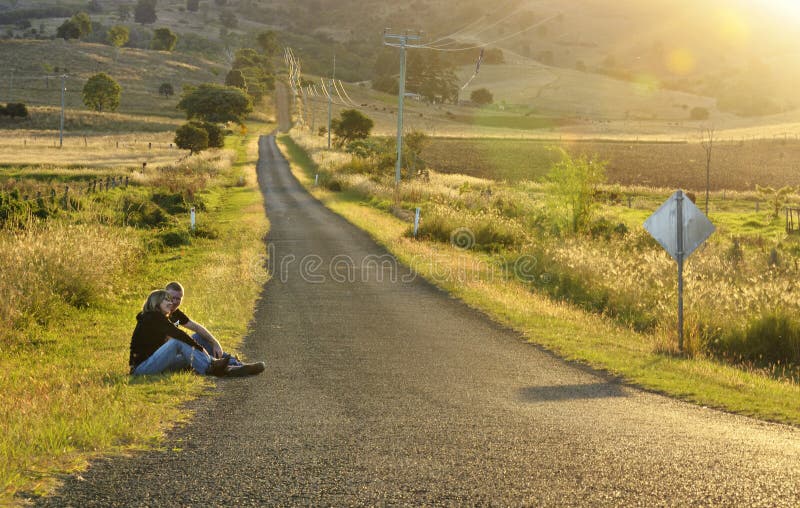 Mutter und Sohn, die durch lange leere aufpassende Sonnenunterganglandschaft der Landstraße sitzen lizenzfreies stockfoto Mutter und Sohn, die durch lange leere aufpassende Sonnenunterganglandschaft der Landstraße sitzen lizenzfreies stockfoto