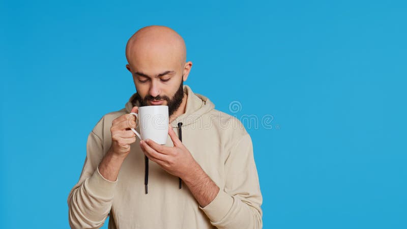 Muslim person enjoying fresh caffeine aroma in studio, drinking cup of coffee and standing over blue background. Middle eastern adult smelling beverage and taking a sip. Camera 2. Handheld shot. Muslim person enjoying fresh caffeine aroma in studio, drinking cup of coffee and standing over blue background. Middle eastern adult smelling beverage and taking a sip. Camera 2. Handheld shot.