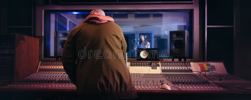 Sound engineer working at audio control panel with singer singing in recording room in background. Musicians producing music in professional recording studio. Sound engineer working at audio control panel with singer singing in recording room in background. Musicians producing music in professional recording studio.