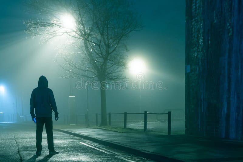 A mysterious hooded figure holding a phone, standing in a city street. On a moody foggy winters night