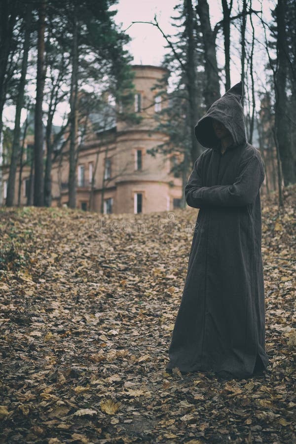 Mysterious monk, wizard or adept of secret society in hooded robe stands in forest against the background of medieval castle or